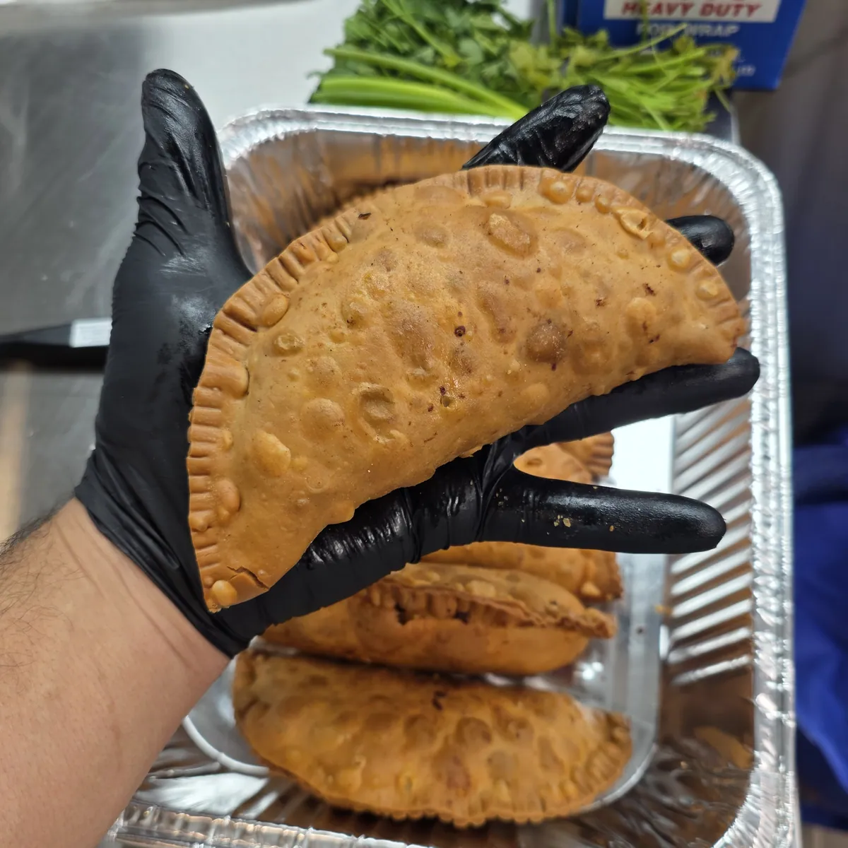 A black-gloved hand holding up a golden Big Papa’s empanada over a tray of fresh empanadas