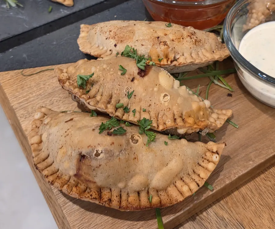 Close-up of crimped golden empanadas on a wood board with parsley and dipping sauces