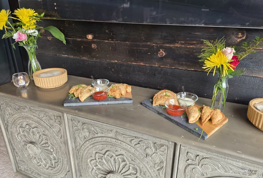 A catering spread of empanada boards with sauces and fresh flowers on a carved sideboard